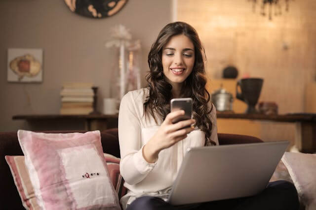 Eine Frau mit langen braunen Haaren sitzt auf einem Sofa, lächelt auf ihr Smartphone und hält einen Laptop auf dem Schoß - vielleicht sucht sie nach Lösungen für Hautprobleme oder bucht einen Termin im Kosmetik ATELIER, einem renommierten Kosmetikstudio in Hamburg. - KOSMETIK ATELIER Hamburg Winterhude